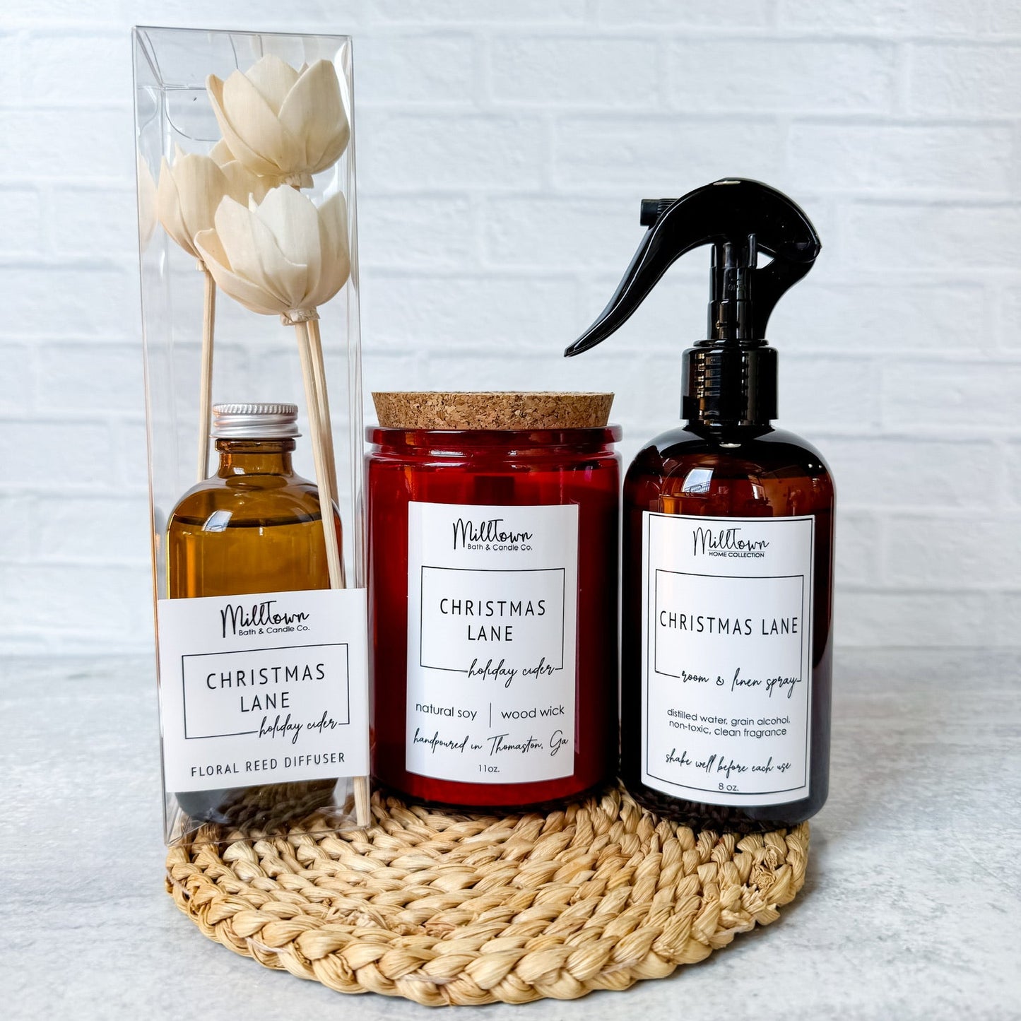 Three MillTown products including a diffuser, candle, and spray bottle on a woven mat with a white brick wall background. Christmas gift Thomaston, GA